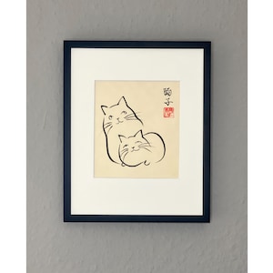Könnte beinhalten: Ein schwarzer gerahmter Druck einer einfachen Strichzeichnung von zwei Katzen, die übereinander liegen. Die Zeichnung ist in schwarzer Tinte auf cremefarbenem Hintergrund. Der Druck hat einen roten Stempel in der oberen rechten Ecke mit dem Text "猫" in Schwarz.