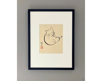 May include: Black framed art print featuring a minimalist line drawing of a cat in a relaxed pose. The artwork is in black ink on a beige background, with Japanese characters and a red stamp. The print is matted with a white border.