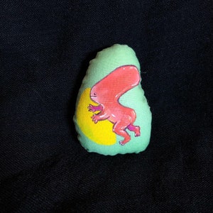 Puede incluir: Una almohada de tela verde en forma de aguacate con una criatura de dibujos animados rosa en su interior. La criatura tiene una luna creciente amarilla detrás.