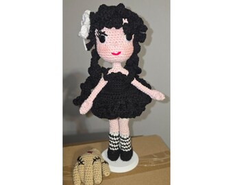 May include: Crocheted doll with black hair, a black dress, and a white bow. The doll has pink skin and is standing on a white base. A crocheted hand with black stitching is next to the doll.