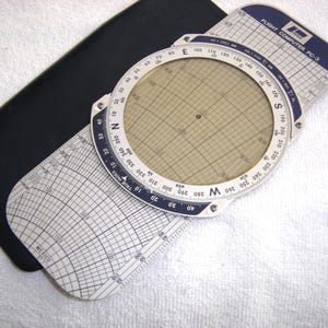 May include: A vintage Flight Computer PC-3, a circular slide rule with a rotating dial and grid, is displayed. The device has a white and blue color scheme with black accents and is set on a black case. The text "FLIGHT COMPUTER PC-3" is visible.