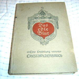 May include: Vintage book titled "Das edle Blut" with a red heart-shaped emblem on the cover. The cover is a light beige color with text in German. The book appears to be old and well-loved.