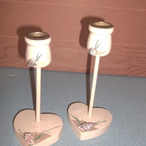 May include: Two white wooden candle holders with heart-shaped bases. Each holder features a seashell and green grass design.