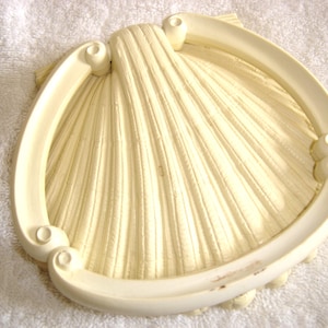 May include: A white ceramic seashell wall decor with a raised, ribbed texture. The shell is framed by a white, decorative border.