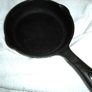 May include: A black cast iron skillet with a long handle. The skillet is on a white towel.