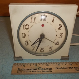 May include: A white vintage Telex electric alarm clock with a round face and black hands. The clock has a light blue trim around the face and a red dot at the 12 o'clock position. The clock is sitting on a blue and white patterned surface.