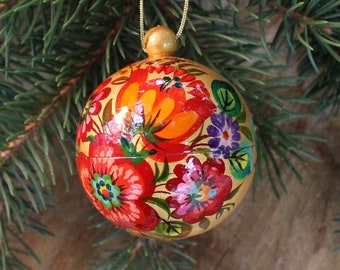 May include: A spherical wooden ornament with a glossy finish, featuring hand-painted floral patterns in vibrant shades of red, orange, purple, and green on a gold background. The ornament is topped with a small gold cap and a thin gold hanging cord.