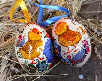 Könnte beinhalten: Zwei bemalte Ostereier aus Holz mit handgemalten Küken und Blumenmustern. Die Eier sind weiß mit gelben, orangefarbenen, blauen, roten und grünen Akzenten.