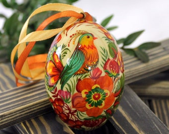 May include: A hand-painted wooden egg ornament with a bird and floral design. The egg is decorated with bright colors, including red, orange, yellow, green, and blue. The ornament is attached to a ribbon.
