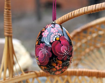 May include: A hand-painted, decorative egg with a glossy black finish. The egg features a floral design in shades of red, pink, and white, with orange and yellow accents. A purple ribbon is attached for hanging. The egg is hanging from a wicker chair.