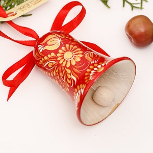 May include: A red wooden bell with a gold floral design and a white interior. The bell is tied with a red ribbon and has a wooden clapper.