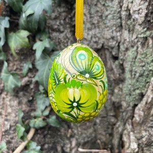 Könnte beinhalten: Ein handbemaltes, grünes und weißes Holzeier-Ornament mit Schmetterlings- und Blumenmuster. Das Ei hängt an einem gelben Band.