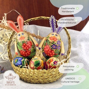 May include: Two hand-painted wooden bunny figurines with floral and berry designs, along with four painted Easter eggs, all in a woven basket. The bunnies are traditional Ukrainian folk art, known as Petrykiwka painting, and are a UNESCO Cultural Heritage.