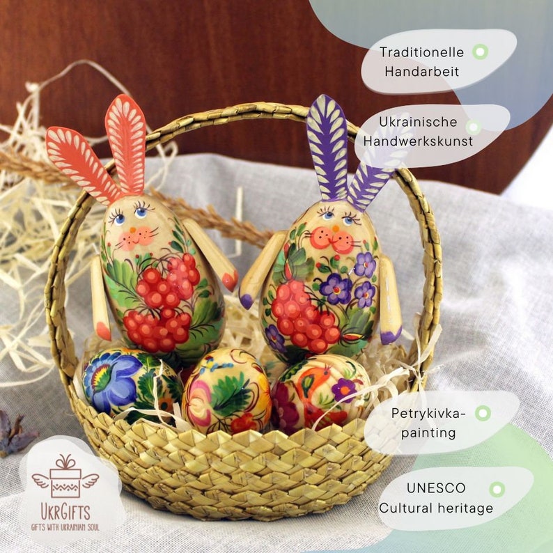 May include: Two hand-painted wooden bunny figurines with floral and berry designs, along with four painted Easter eggs, all in a woven basket. The bunnies are traditional Ukrainian folk art, known as Petrykiwka painting, and are a UNESCO Cultural Heritage.
