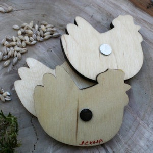 May include: Two wooden chicken shaped magnets with a natural wood finish. The magnets are attached to the back of the chickens. The chickens are on a wooden surface.