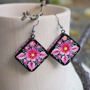 May include: A pair of black square earrings with pink and white floral designs. The earrings have silver hooks.