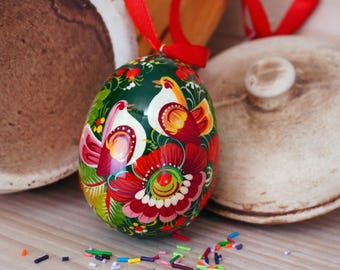 Könnte beinhalten: Ein grünes Osterei mit einem bemalten Design aus roten und gelben Blumen, weißen Vögeln und grünen Blättern. Das Ei hängt an einem roten Band.