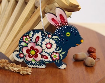 Könnte beinhalten: Ein blauer Holz-Hasen-Anhänger mit rosa Ohren und weißen Blumen, die auf seinen Körper gemalt sind. Der Hase hat ein weißes und schwarzes Auge und ein weißes Punktmuster auf seinem Körper.