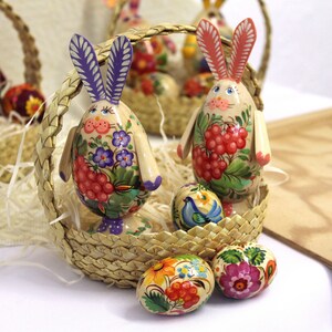 May include: A wicker basket filled with colorful, hand-painted Easter eggs and wooden bunny figurines. The eggs are decorated with floral patterns and the bunnies have intricate details.