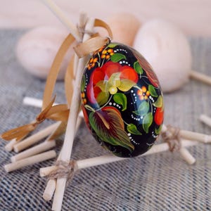 Puede incluir: Un huevo de Pascua pintado a mano con fondo negro y diseños florales coloridos. El huevo está en un soporte de madera.