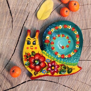 Pu&ograve; includere: Una lumaca di legno dipinta a mano con un guscio giallo, una spirale verde e blu e fiori rossi e gialli. La lumaca &egrave; su una superficie di legno.