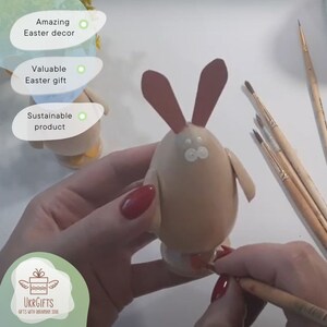 May include: A hand holds a brown egg painted to look like a bunny. The bunny has red ears, white eyes, and a red nose. The egg is sitting on a white surface. There are paintbrushes in the background.