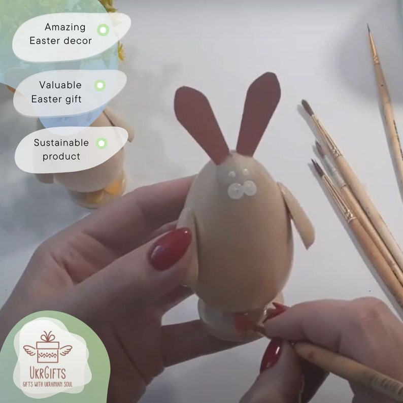 May include: A hand holds a brown egg painted to look like a bunny. The bunny has red ears, white eyes, and a red nose. The egg is sitting on a white surface. There are paintbrushes in the background.