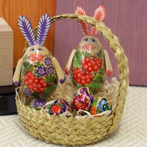 May include: Two hand-painted wooden bunny figurines with floral and berry designs, sitting in a woven basket with four painted Easter eggs.