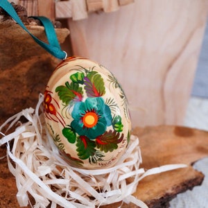 May include: A wooden egg ornament with a hand-painted floral design in shades of green, blue, yellow, and red. The egg is hanging from a teal ribbon and is sitting on a bed of shredded paper.