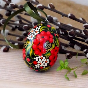Puede incluir: Un huevo de madera negro con un diseño pintado a mano de bayas rojas y flores blancas. El huevo está decorado con detalles dorados y tiene una cinta verde.