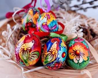Könnte beinhalten: Fünf handbemalte Ostereier mit bunten Blumen- und Vogelmotiven. Die Eier sind in einem Korb mit Stroh und einem Weidenzweig angeordnet.