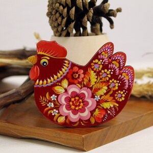 May include: A red wooden chicken figurine with a floral design painted on it. The chicken has a yellow beak and black eyes. The flowers are painted in shades of pink, yellow, orange, and green.