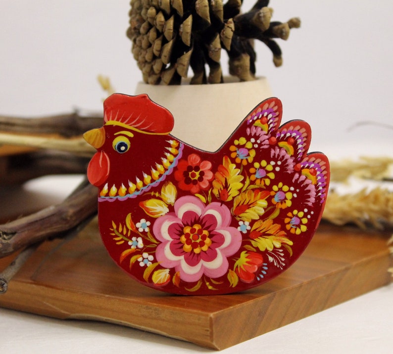 May include: A red wooden chicken figurine with a floral design painted on it. The chicken has a yellow beak and black eyes. The flowers are painted in shades of pink, yellow, orange, and green.