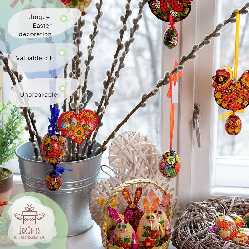 May include: A collection of hand-painted wooden Easter eggs, some with floral designs, others with bunny faces, and one shaped like a heart. The eggs are displayed in a wicker basket and a metal bucket, with willow branches and a white heart-shaped decoration in the background.