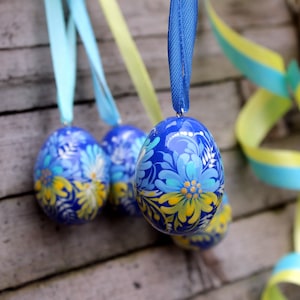 Puede incluir: Adornos de huevos de Pascua azules pintados a mano con diseños florales en azul, amarillo y blanco. Cada huevo está suspendido por una cinta azul. El fondo presenta una superficie de madera y cintas decorativas.