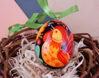 Könnte beinhalten: Ein handbemaltes Osterei mit schwarzem Hintergrund und einem bunten Hahn-Design. Das Ei ist mit roter, gelber, orangefarbener, grüner und blauer Farbe verziert. Das Ei liegt in einem Nest aus Zweigen und Stroh.
