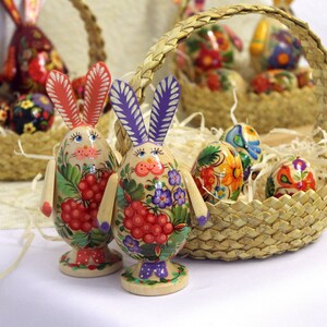 May include: Two hand-painted wooden Easter bunny figurines with floral and berry designs. The bunnies are standing on a white surface in front of a wicker basket filled with colorful Easter eggs.