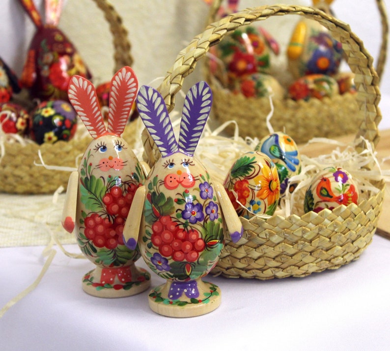 May include: Two hand-painted wooden Easter bunny figurines with floral and berry designs. The bunnies are standing on a white surface in front of a wicker basket filled with colorful Easter eggs.