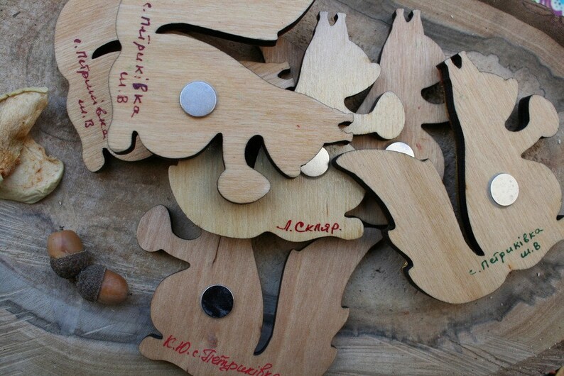 Pu&ograve; includere: Un set di magneti in legno a forma di scoiattolo con il testo "с. Петриківка ш. В" e "Л. Скляр." scritto su di essi.