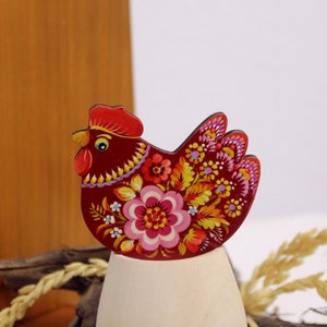May include: A red wooden chicken figurine with a floral design painted on it. The chicken is standing on a white egg-shaped base.