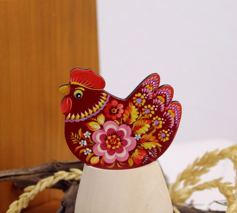 May include: A red wooden chicken figurine with a floral design painted on it. The chicken is standing on a white egg-shaped base.