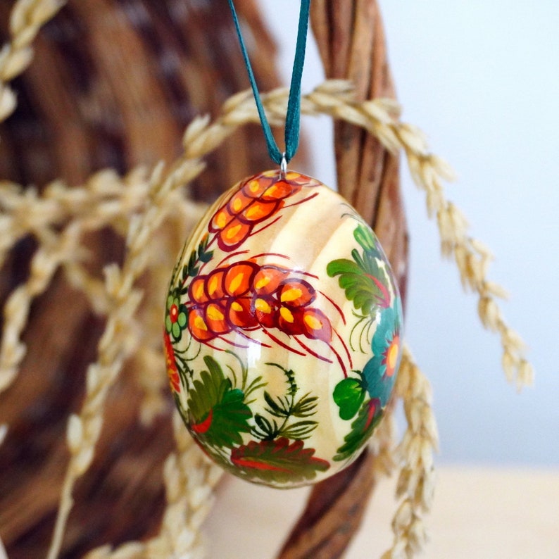 May include: A hand-painted wooden egg ornament with a white background and a floral design featuring orange, red, green, and yellow flowers. The egg is hanging from a green ribbon.