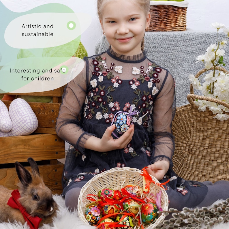 May include: A young girl holds a decorated Easter egg in her hand. A basket filled with colorful, hand-painted Easter eggs sits in front of her. The eggs are decorated with floral patterns and bright colors. The girl is wearing a black dress with floral embroidery. The image is set against a backdrop of a wooden crate, a wicker basket, and a white fluffy surface.