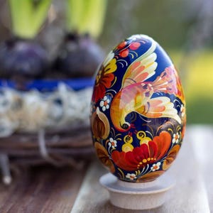 Op de afbeelding: Een handbeschilderd paasei met een blauwe achtergrond en een gele vogel met oranje vleugels. Het ei is versierd met rode, oranje en gele bloemen.