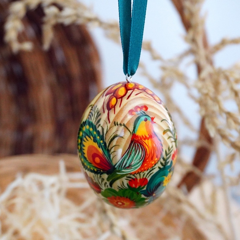 May include: A wooden Easter egg ornament with a hand-painted design of a colorful bird with a long tail and flowers. The egg is hanging from a teal ribbon.