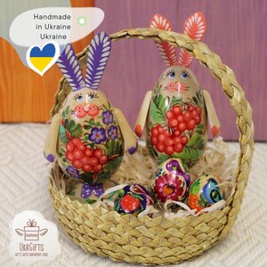 May include: Two hand-painted wooden Easter bunny figurines with floral designs and red berries, along with four smaller painted eggs, all in a woven basket. Handmade in Ukraine.
