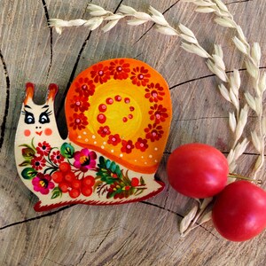 Pu&ograve; includere: Una lumaca di legno dipinta con fiori rossi, gialli e verdi brillanti. La lumaca &egrave; su una superficie di legno con due prugne rosse.