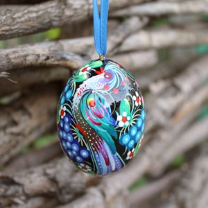 Könnte beinhalten: Ein schwarzer, eiförmiger Anhänger mit einem bunten, bemalten Design eines Vogels mit blauen, grünen, rosa und gelben Federn. Der Anhänger ist mit weißen Blumen und blauen Trauben verziert.