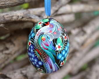 May include: A black egg-shaped ornament with a colorful painted design of a bird with blue, green, pink, and yellow feathers. The ornament is decorated with white flowers and blue grapes.