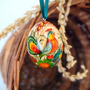 May include: A hand-painted wooden Easter egg with a colorful rooster design. The egg is decorated with flowers and leaves in shades of red, green, yellow, and orange.